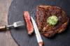 Medium gebratenes Steak mit Kräuterbutter, mit Messer und Gabel angeschnitten auf neumodischer, runder Schieferplatte serviert