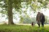 Paar läuft Arm in Arm mit Gummistiefeln und mit Hund über eine Wiese mit Eichenbaum