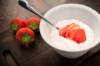 Eine weisse Schale mit Quark und einer geschnittenen Erdbeere und Löffel, daneben hat der Fotograf drei ganze frische Erdbeeren drapiert