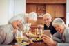 Zwei Rentnerpäarchen sitzen am Tisch vor Schrankwand, essen und trinken Wein, eine Frau zeigt Fotos auf ihrem Smartphone, die alle belustigen