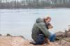 Mann in Parka sitzt am See und liebkost seinen Hund in einer sehr intimen Situation